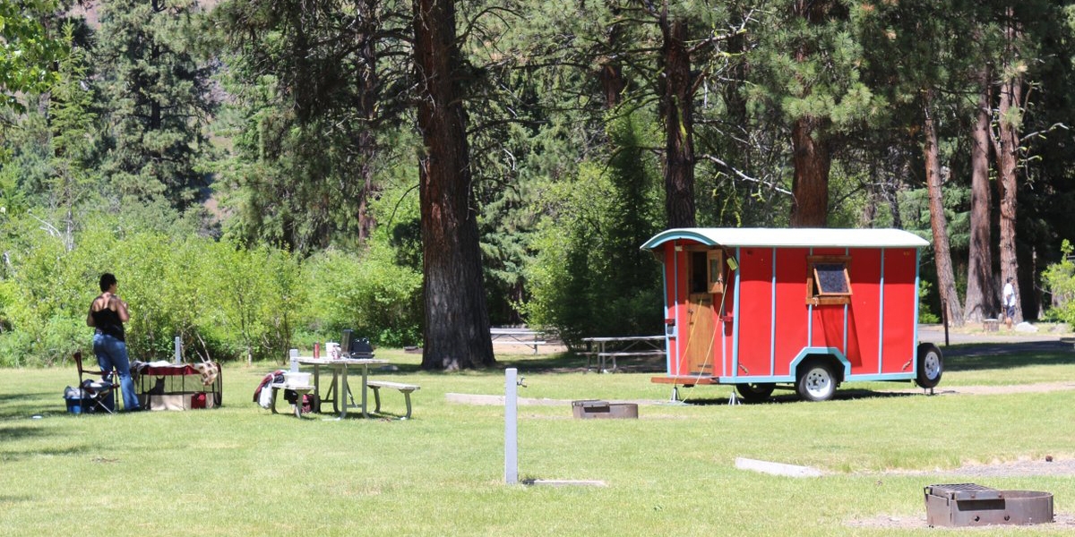 A red trailer is parked in a campsite, with fire pit, picnic table, and water spigot in view. A person stands near the picnic table.